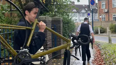 Alasdair is holding his bike outside, it is mustard yellow in colour and does not have any wheels. There is a policeman holding another bike ahead.