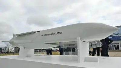 A Storm shadow missile stands against the backdrop of some grey clouds with people wandering around looking at it from the opposite side of the camera.,