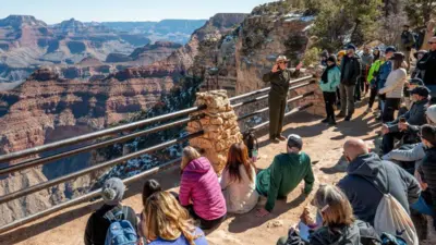 A group of visitors in brightly coloured sportswear sit on the ground or stand to listen to a Grand Canyon tour guide, in a park ranger hat and olive uniform, who is standing facing them at the railing at the edge of the canyon. Beyond the railing is the sepia coloured canyon