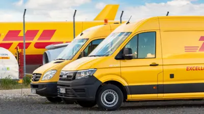 Dwa żółte samochody dostawcze DHL stoją przed samolotem transportowym zaparkowanym na terminalu lotniska Lipsk-Halle