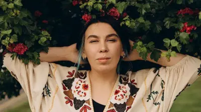 Nastya Zukhvala next to a leafy bush with red flower looking directly into the camera. 