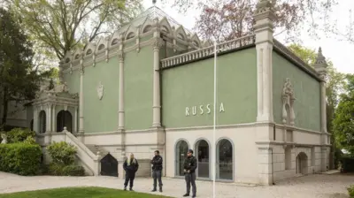 Büyük, iki- üç katlı yeşil bir bina ve üzerinde latin harfleriyle Rusya yazıyor. Kapısında üç güvenlik görevlisi duruyor.