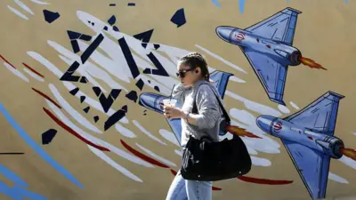 یک زن جوان ایرانی از کنار نقاشی‌های دیواری رنگارنگ عبور می‌کند، در حالی که زندگی روزمره در سایه ویرانی ناشی از حملات آمریکا و اسرائیل بر تهران، ایران، در تاریخ ۱۶ فروردین ۱۴۰۵ ادامه دارد. با وجود حملاتی که از ۹ اسفند ۱۴۰۴ آغاز شد و منجر به کشته شدن رهبر ایران، آیت‌الله علی خامنه‌ای، و شمار زیادی از غیرنظامیان شد، مردم محلی در خیابان‌هایی با ساختمان‌ها و خودروهای آسیب‌دیده رفت‌وآمد می‌کنند و در تلاشند تا حدی حس عادی بودن زندگی را حفظ کنند.