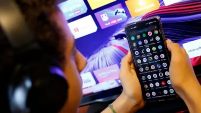 Adolescente em frente a um computador segura um celular com vários aplicativos aparecendo na tela. 