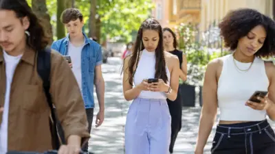 Grupa osób idzie ulicą, patrząc na ekrany telefonów komórkowych. Jest ciepły, słoneczny dzień. 