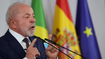 O presidente Lula, de barba e cabelo grisalhos, veste terno escuro, camisa branca e gravata, enquanto discursa em um púlpito com dois microfones à frente. Ele gesticula com as mãos levantadas, apontando os dedos indicadores. Ao fundo, aparecem bandeiras desfocadas — do Brasil, da Espanha e da União Europeia.