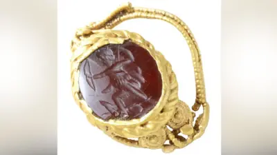 The front of a gold Roman ring, with a red stone carved with a view of Diana pulling back her bow set in its middle, surrounded by its gold setting. The gold of its crumpled setting and band is ornately worked 