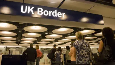 UK airport passport control. There is a blue overhead sign with UK border written on it in white. People are queuing to get their passports checked.