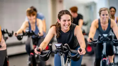 Una mujer hace ejercicios en una bicicleta estática mientras sonríe. Personas en el fondo también hacen ejercicio en bicicletas estáticas. Se ven fuera de foco. 