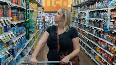 Nicole Williams, a woman with blonde hair in a black t-shirt, is shopping at a grocery store in Las Vegas, Nevada. She is pushing a trolley down an aisle and looking at cereal bars.