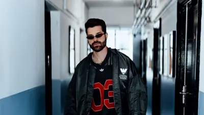 Alok, um homem de barba e cabelo escuro, posa em um corredor de paredes brancas e azul-acinzentadas, olhando diretamente para a câmera com expressão séria. Ele veste um casaco oversized preto sobre uma camisa esportiva preta com o número "03" em vermelho, além de óculos de sol estreitos e um colar prateado, compondo um visual de inspiração urbana e contemporânea.