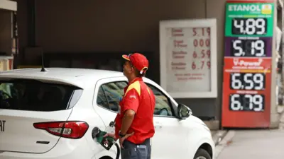 Um frentista abastece um carro branco em um posto de gasolina com uma placa de preços ao fundo.