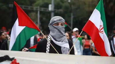 یک تظاهرکننده حامی فلسطین و جمهوری اسلامی که چهره‌اش را پوشانده، دو پرچم جمهوری اسلامی و فلسطین را در دست گرفته است