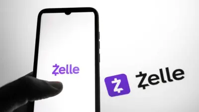 Uma mão segurando um celular com o logotipo do aplicativo Zelle na tela, com letras roxas sobre um fundo branco. Ao fundo, é possível ver o mesmo logotipo, nas cores preto e roxo