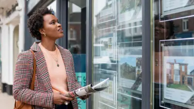 A woman wearing a checked jacket and holding a brochure looks in an estate agent's window