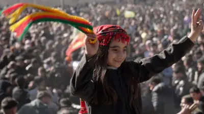 A child in black top and traditional read scarf on her head, waves a green, red and yellow scarf as she sits on someone's shoulders at anniversary celebrations.  A large crowd can be seen behind her. 