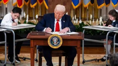 Donald Trump sits at a desk signing an executive order. He is surrounded by children sitting at desks writing on paper