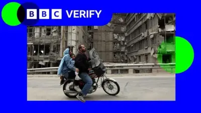 یک مرد و زن و کودکی خردسال سوار بر موتور از مقابل ساختمان‌های آوار عبور می‌کنند، لوگوی بی‌بی‌سی وریفای در گوشه تصویر دیده می‌شود