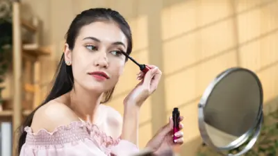 Woman with long dark hair, wearing a pink off-the-shoulder top looking in a small mirror while she applies mascara