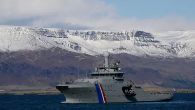 Un barco de los guardacostas de Islandia, en el agua, con un paisaje de montañas nevadas al fondo. 