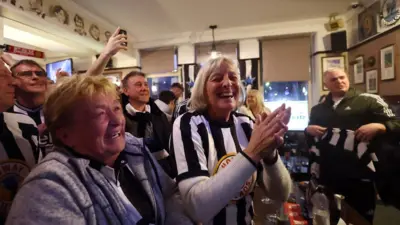 Group of people in a pub - most sporting Newcastle United strips or scarves - with the main focus on two women smiling and crying at the same time.