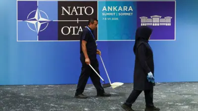 NATO yazan mavi panonun önünde iki temizlik görevlisi