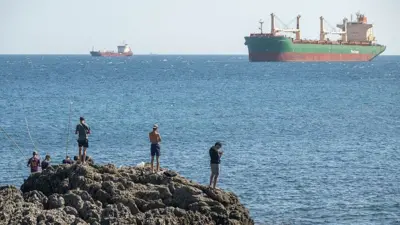 Bir grup erkek, arkadan görülüyor; İspanya'nın Ceuta kentinde, 22 Ağustos 2026 tarihinde kayalık bir çıkıntı üzerinde, arka planda mavi deniz ve büyük gemiler yer alıyor.