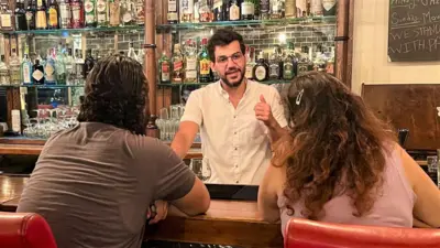 Bartender with bottles of liquor behind him, speaking to two customers across the bar 