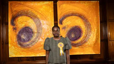 تصویر ننا کالو، هنرمند، که مقابل دو اثر طراحی خود با رنگ‌های زرد و بنفش ایستاده است