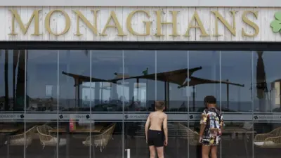 Two boys peering inside Monaghans bar in Costa del Sol. One of them is wearing black shorts and the other is wearing a long shirt covering his shorts.