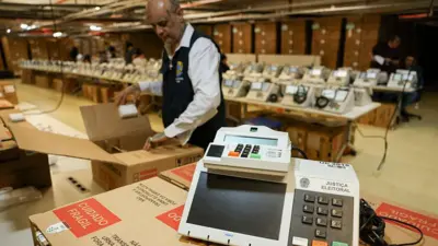 urna eletrônica sobre uma caixa. No fundo, várias urnas e um funcionário da Justiça Eleitoral. 