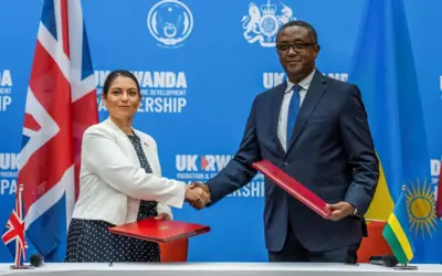 Umutegetsi w'umugore uhagarariye Ubwongereza n'umutegetsi w'umugabo uhagarariye u Rwanda bahagaze bahanye ikiganza, nyuma yo gusinya amasezerano ku bimukira mu 2022. Inyuma ku ruhande hari ibendera rinini ry'Ubwongereza n'iry'u Rwanda, imbere ku meza hari amabendera abiri mato nk'ayo.