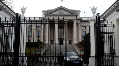 Ambasada Rusiei din Polonia - o clădire neoclasică impunătoare, care are o poartă masivă din fier forjat în față. O mașină neagră se vede trecând prin poarta deschisă. 