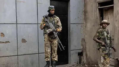 Abagabo babiri bo mu nyeshyamba za M23 bambaye imyenda ya gisirikare bahagaze bafite imbunda nini, i Goma, mu burasirazuba bwa Repubulika ya Demokarasi ya Congo, ku itariki ya 23 Gashyantare (2) mu 2025. Inyuma yabo hari inkuta z’inzu.