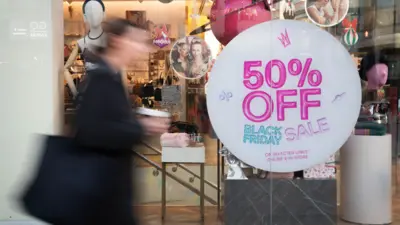 An out-of-focus person walks in front of a shop window with a sign saying '50% off Black Friday sale', taken on Oxford Street in November 2023