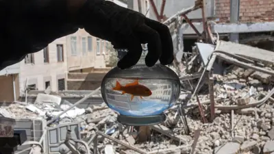 تهران، ایران – دهم فروردین: مردی یک ماهی قرمز را در تنگ آن در برابر آپارتمان آسیب دیده در حمله هوایی نشان می‌دهد