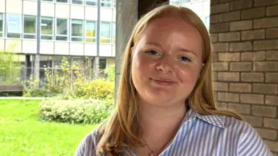 A head-and-shoulders shot of Isobel, outside her office at work, smiling into the camera. She has long, strawberry-blonde hair and wears a striped blue and white shirt.