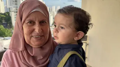 Senhora com véu islâmico e bebê no colo; ao fundo, prédios de São Paulo