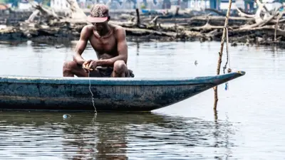 One man sit down for boat e dey hold fishing line for di rive in Niger Delta. Behind am ypu fit see dead mangroves and some blurry buildings.