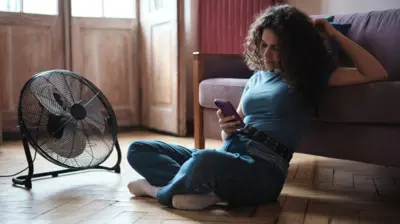 Egy fiatal nő a telefonját görgeti, a padlón ülve, egy ventilátor előtt
