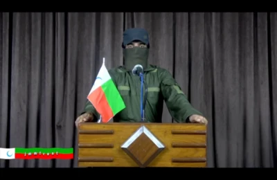 تصویری برگرفته از ویدیویی که برای اعلام تشکیل «جبهه مبارزین مردمی» منتشر شد و سخنگوی نقاب‌دار گروه تازه در آن یک تریبون در حال صحبت است. جلوی او یک میکروفون و طرف چپ میکروفون یک پرچم به رنگ‌های سبز، قرمز و سفید با حلالی در بخش سفید آن دیده می‌شود.