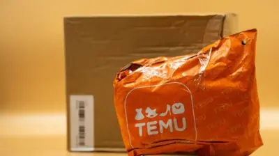 Turuncu renkte bir duvarın önünde duran bir TEMU kolisi ve poşeti. 