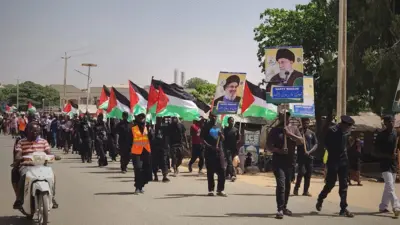 Ndị na-eme ngagharịiwe na-aga ije n'ahịrị ma yiri uwe oji ma jiri ọkọlọtọ mba Iran nakwa foto onyendu kachasị elu na Iran e gburu egbu, Ayatollah Khamenei. 