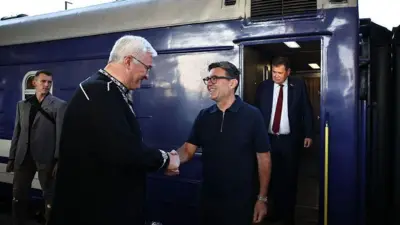 Burnham îi strânge mâna lui Sybiha, în timp ce amândoi zâmbesc, stând alături de alți doi bărbați și lângă un tren.