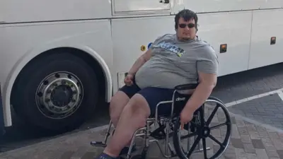 Charley Jonstone-Brent who uses a mobility aid, sitting in a wheelchair in front of a coach. It's a bright day and he is wearing sunglasses, shorts and a t-shirt.