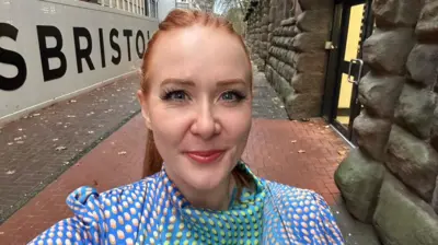 Anna Hill smiling while stood outside. She has ginger hair in a ponytail and is wearing a bright blue dress with dots.