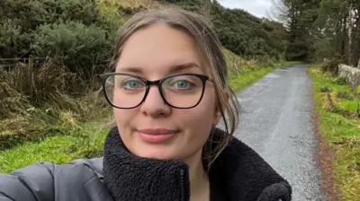 Weronika takes a selfie on a country lane, with a path leading off behind her and hedges on her right side, a row of trees on her left. It's a damp day and she is wearing a black puffer jacket with a fleece lining. She is also wearing stylish large-framed glasses, her light brown hair pulled back in a pony tail.