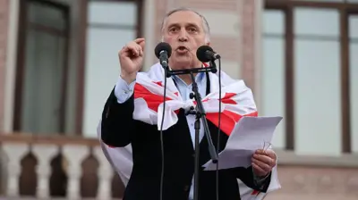 Паата Бурчуладзе, седой мужчина в черном пиджаке с накинутым на плечи грузинским флагом, говорит в два микрофона. В его левой руке бумаги