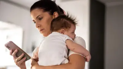 žena sa bebom u naručju drži u ruci mobilni telefon