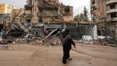 A woman dressed in a black headscarf and dark clothing walks past the wreckage of heavily damaged buildings in Beirut’s southern suburbs, Lebanon. The scene shows extensive destruction, with collapsed walls, twisted metal, and debris scattered across the ground. 